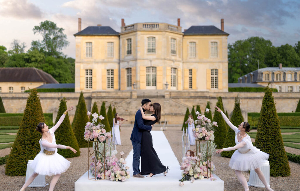 Newly engaged couple kissing on a white platform at Château Villette, two ballet dancers performing beside them, pink and white floral arrangements on gold stands, violinist playing in the background, French château behind