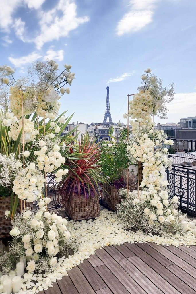 Luxury elopement floral setup on the rooftop terrace of Hôtel Marignan Champs-Élysées in Paris, gold stands with white rose and gypsophila arrangements, white rose petal carpet on wood decking, tropical plants in wicker baskets, Eiffel Tower centered directly behind under a blue sky