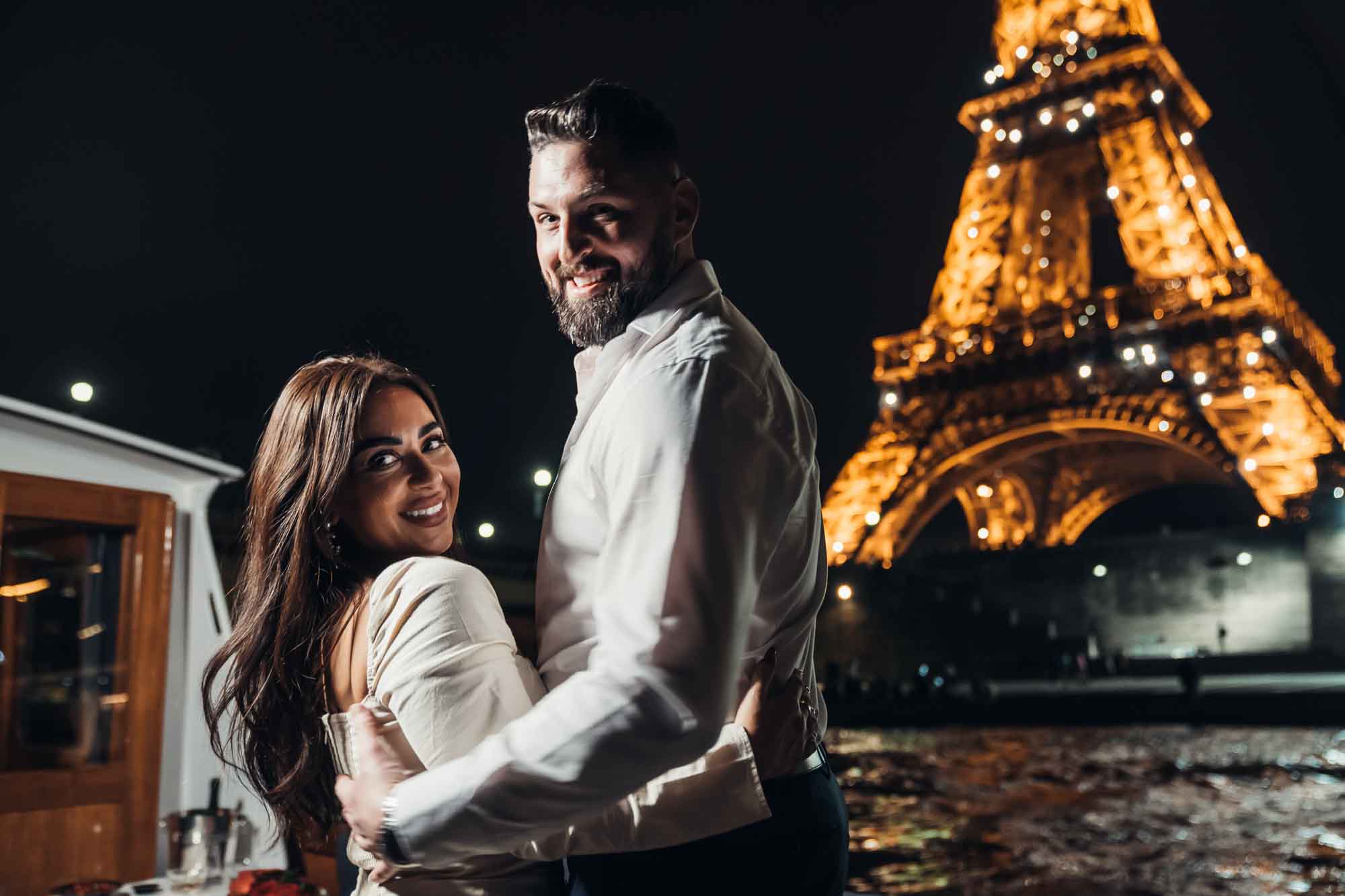Smiling couple embracing on a private yacht at night in Paris, with the sparkling Eiffel Tower in the background during their romantic Seine River cruise.