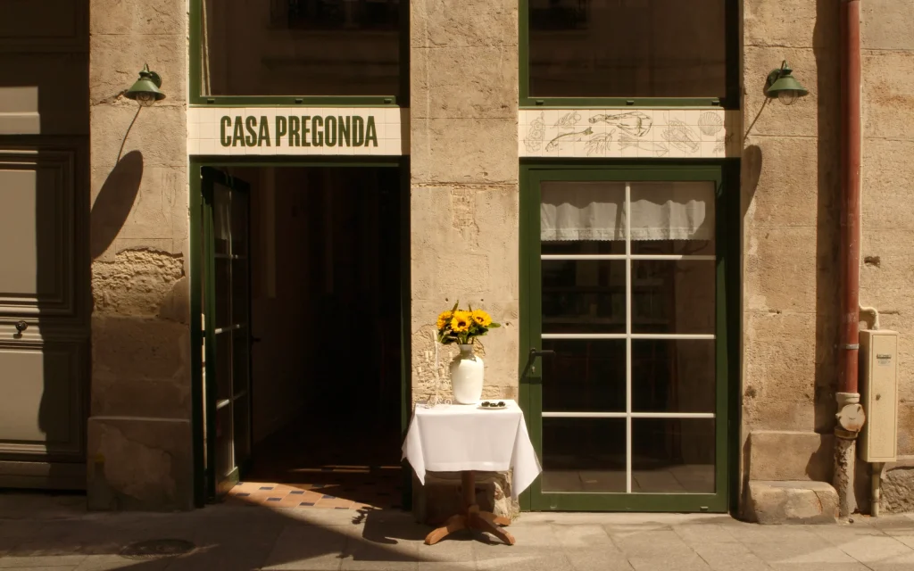 Sun-lit façade of Casa Pregonda restaurant in Paris with green-framed windows and sunflowers at the entrance