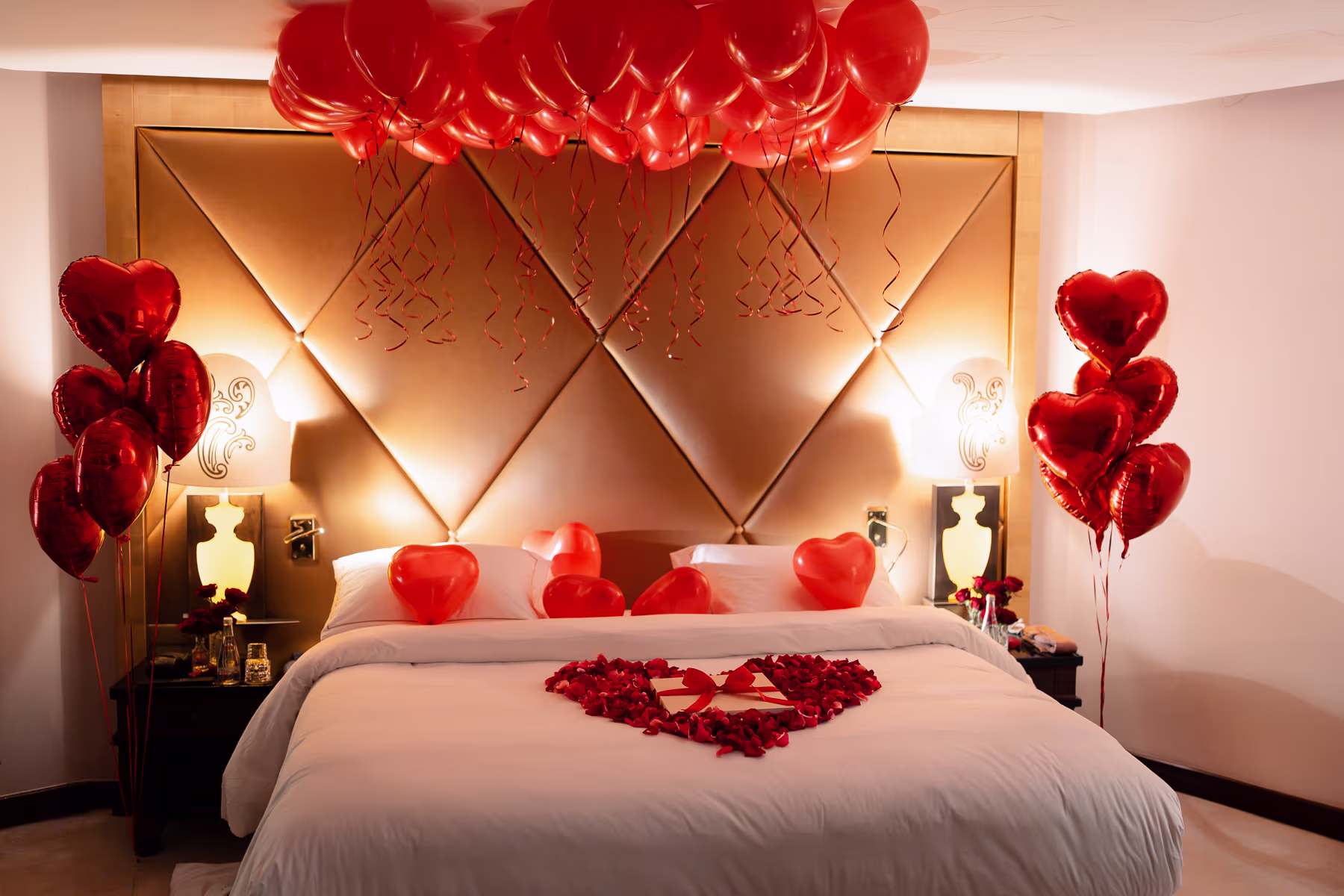 Romantic Paris hotel suite with premium room decor, red heart helium balloons around the bed, rose-petal heart on the duvet framing a gift box, and chilled champagne for a surprise proposal or anniversary
