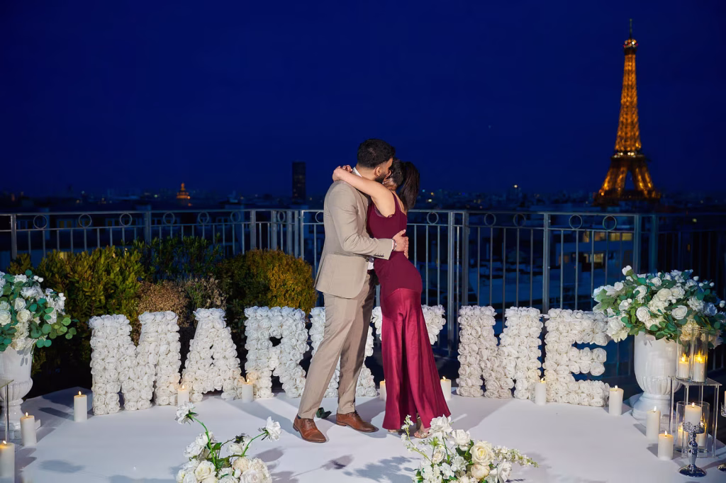 Couple embracing on the Copernic rooftop in Paris at night, large white rose marry me letter display behind them, candles and white floral arrangements, illuminated Eiffel Tower in the background