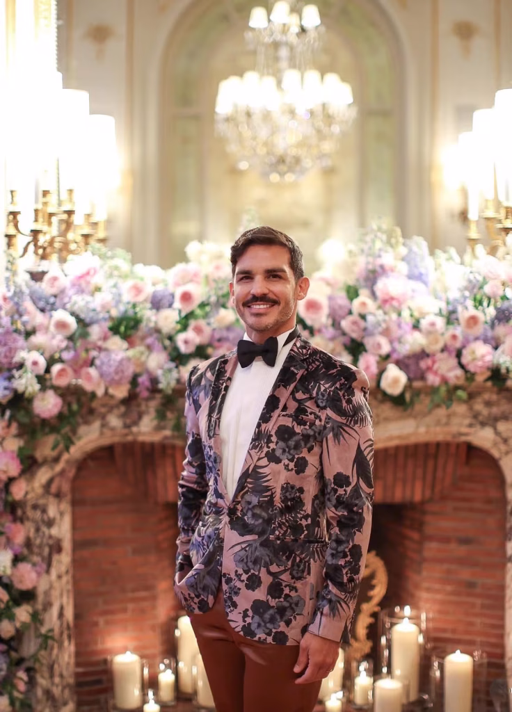 Efren Prieto smiling in a floral tuxedo jacket and bow tie inside an ornate Parisian ballroom with crystal chandeliers, pink and lavender floral arrangements on a stone fireplace and candles behind him