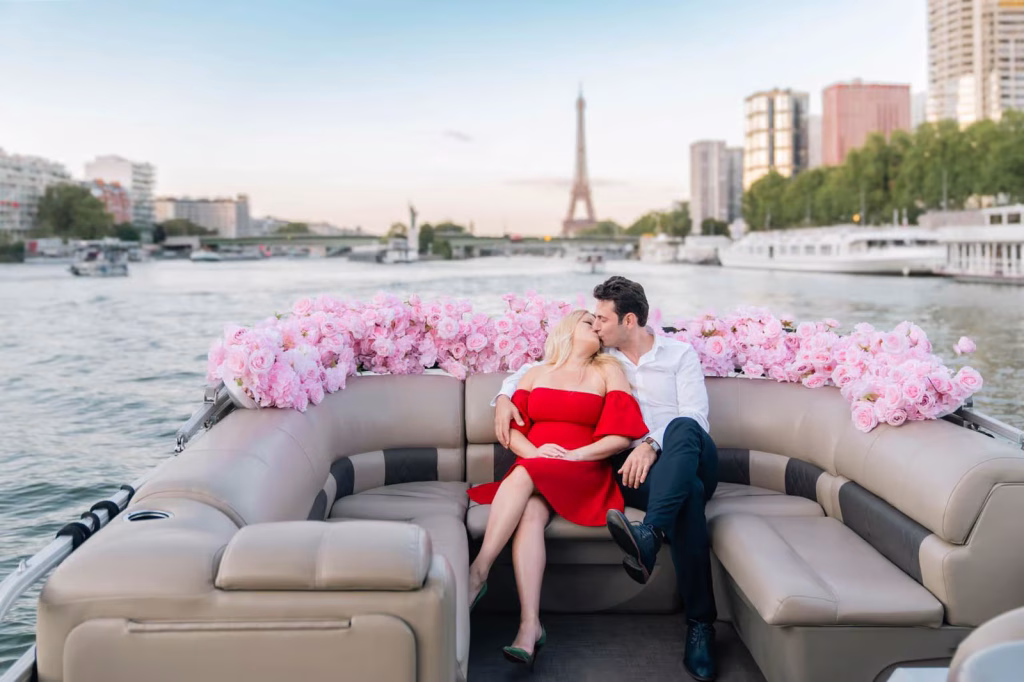 Newly engaged couple kissing on a private boat on the Seine river, surrounded by pink rose arrangements along the railing, Eiffel Tower in the background at sunset