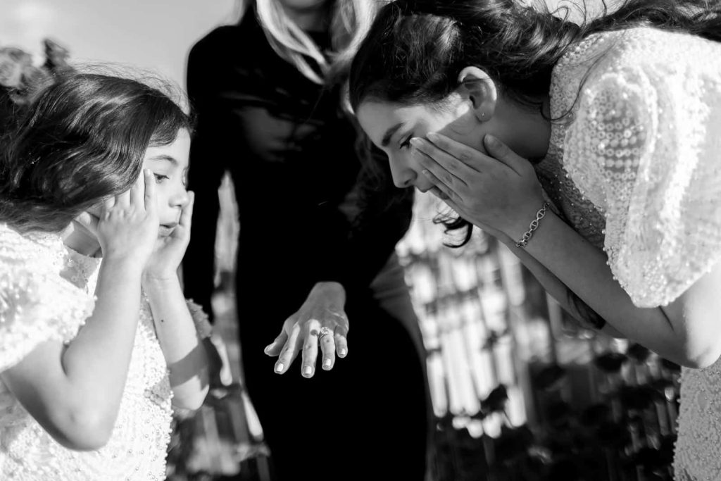 Black and white close-up of newly engaged woman showing her diamond engagement ring to emotional family members after a private rooftop proposal at Le Passy in Paris, two young girls reacting with hands on faces in shock and joy
