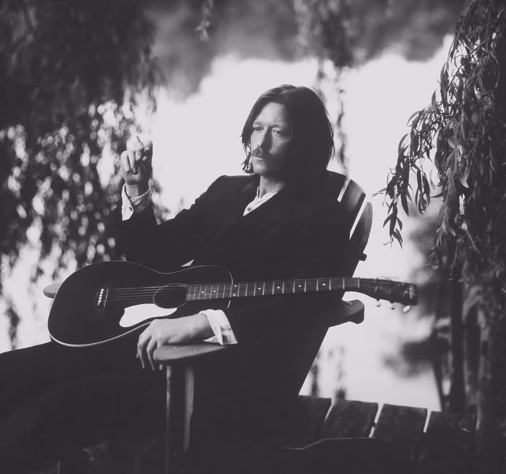 Black and white portrait of Peter Deaves seated with an acoustic guitar outdoors, dark jacket, willow branches framing the shot
