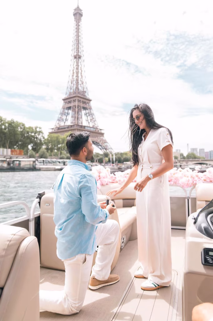 Man on one knee proposing to woman in white jumpsuit on a private boat on the Seine, pink rose arrangements behind them, Eiffel Tower centered in background