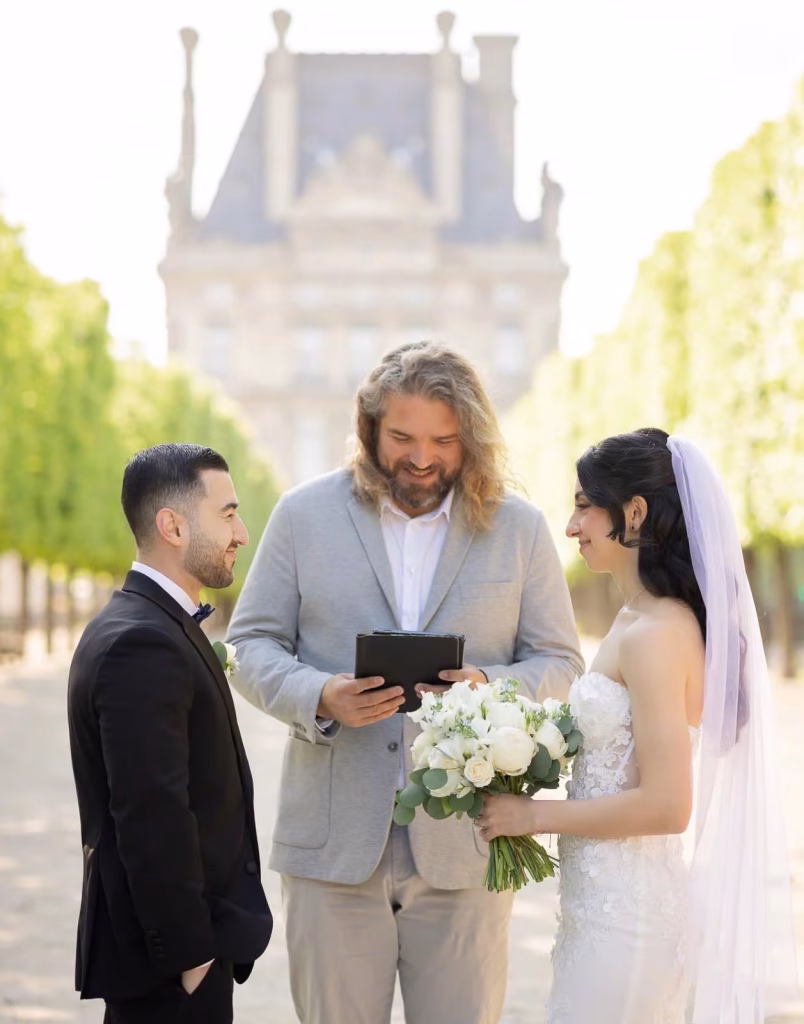 Paris Celebrants officiant Marcus reading from ceremony book between bride in lace gown with white bouquet and groom in black tuxedo during elopement ceremony at Tuileries Garden, Louvre pavilion softly blurred behind