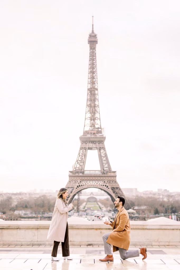 Man on one knee proposing to woman in beige coat on the Trocadéro platform in Paris, Eiffel Tower and Champ de Mars centered directly behind, ring box open